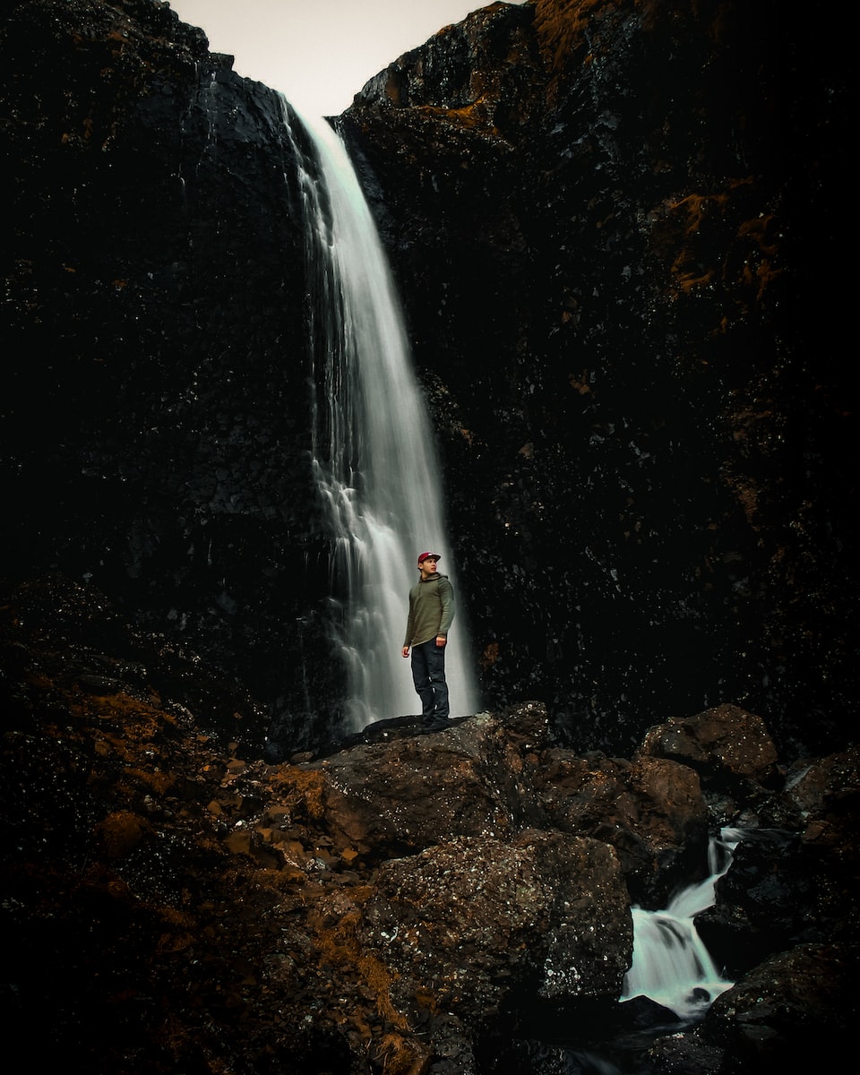 man in white shirt standing on brown rock near waterfalls during daytime